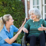 Carer Pushing Senior Woman In Wheelchair Outside Home